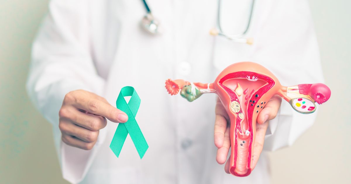A stock photo shows a doctor holding the teal ribbon for ovarian cancer while holding a model of a woman's uterus and ovaries.